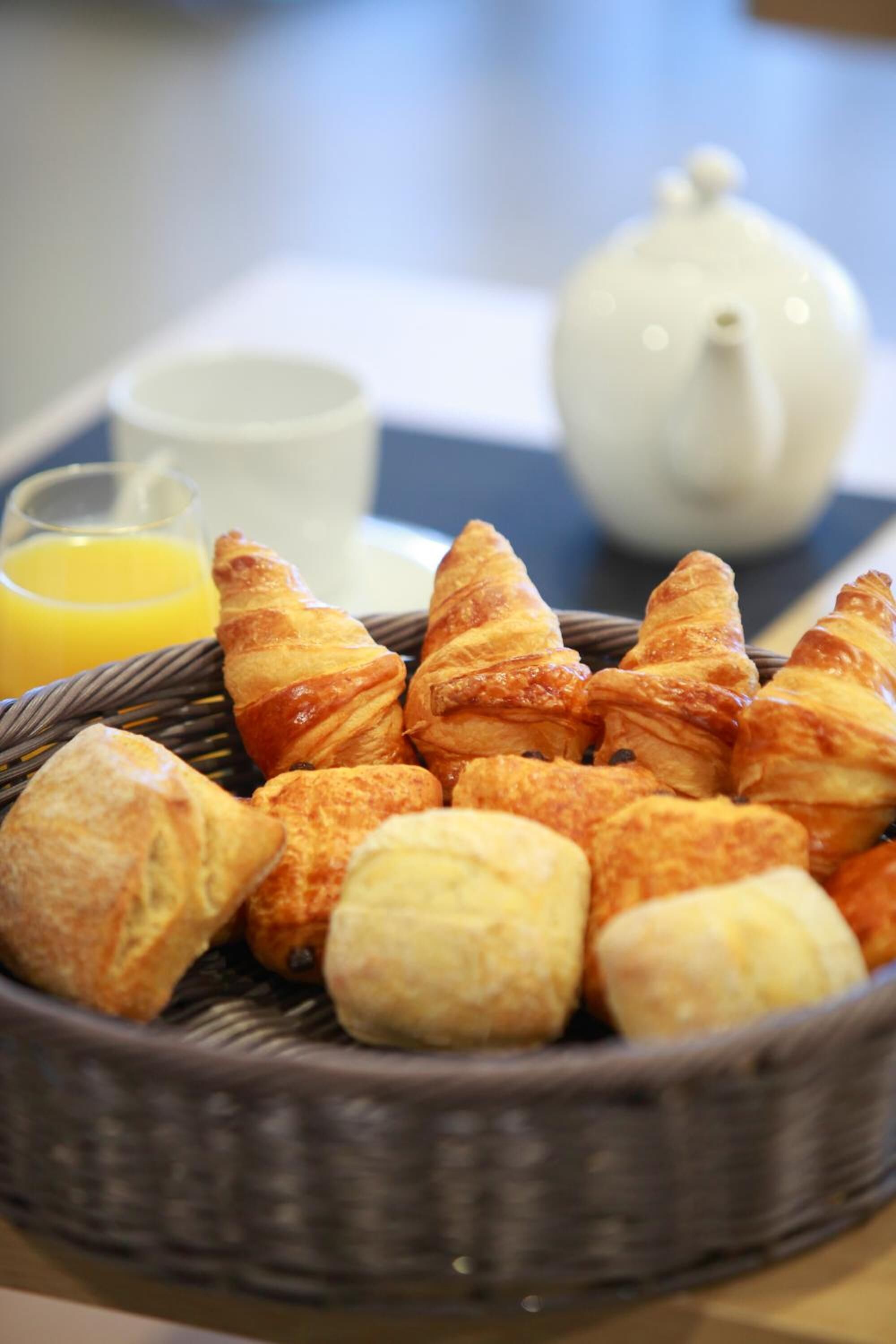 Un panier de croissants et une théière sur un plateau, accompagnés d’un verre de jus d’orange.