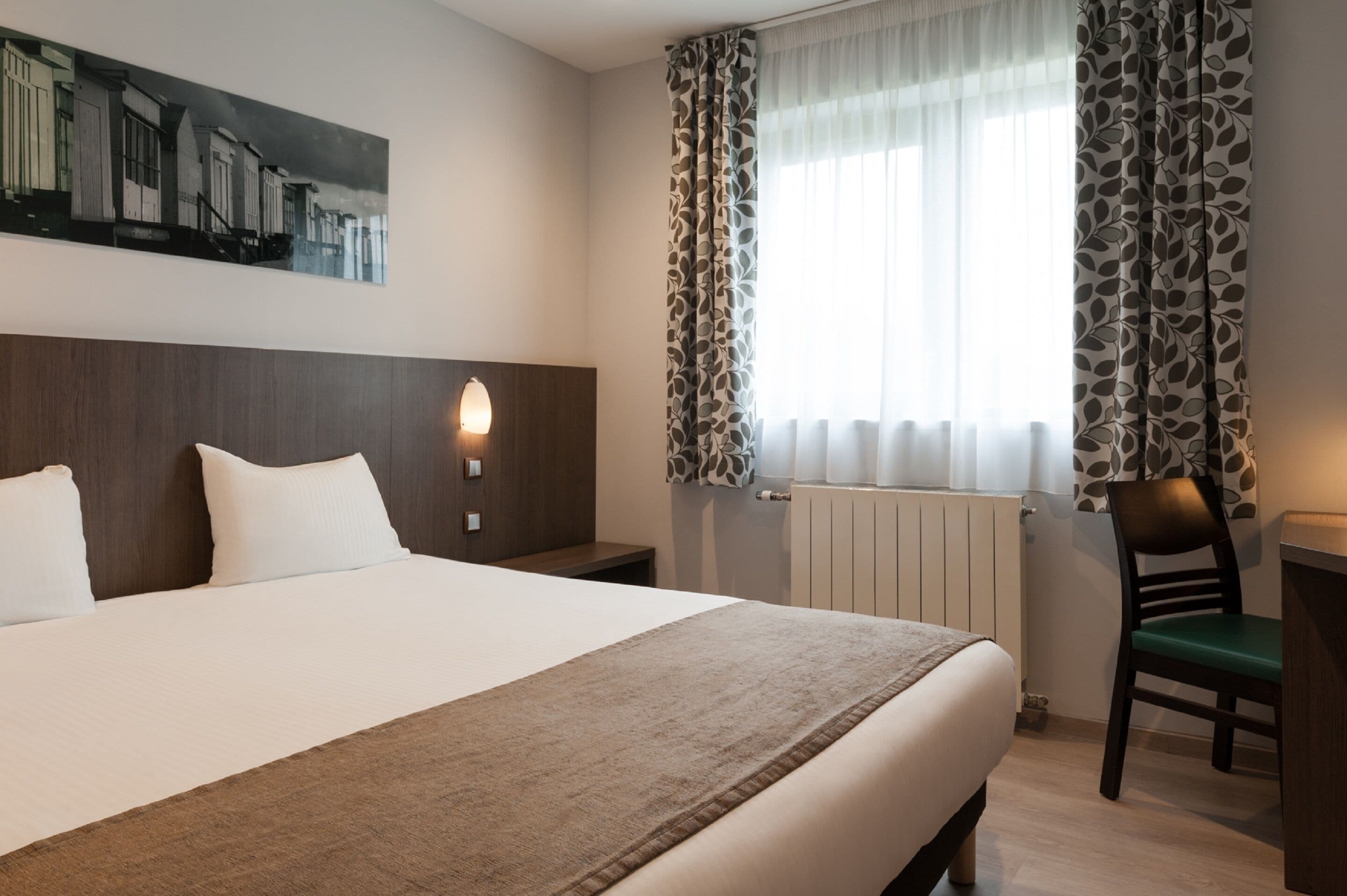 Et hotelværelse med en stor seng, et skrivebord, en stol, et natbord, en vægmonteret lampe og et vindue med mønstrede gardiner.