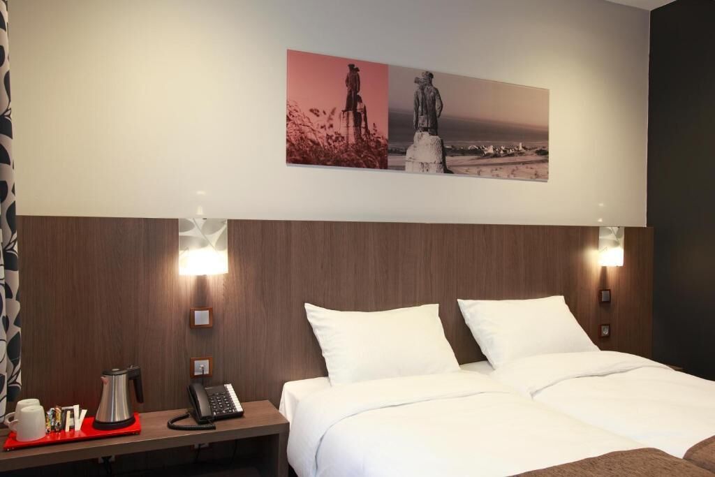 A hotel room with a bed, a nightstand with a phone and a coffee maker, and a wall-mounted picture.
