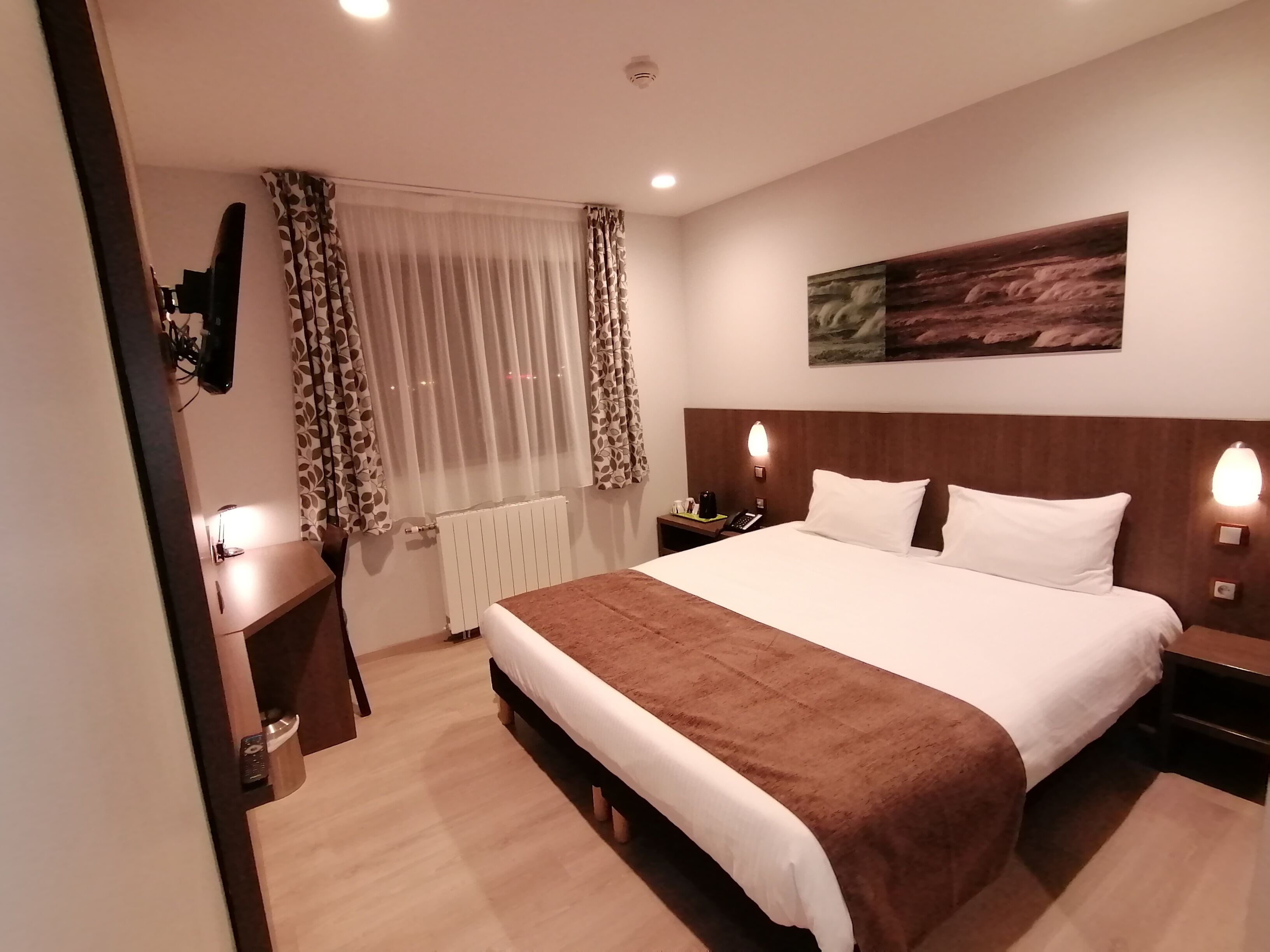 A hotel room with a large bed, bedside tables, a desk, a chair, a radiator, curtains, and a painting on the wall.