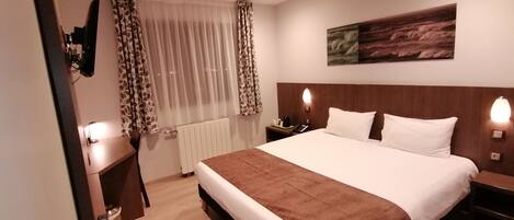 A hotel room with a large bed, bedside tables, a desk, a chair, a radiator, curtains, and a painting on the wall.