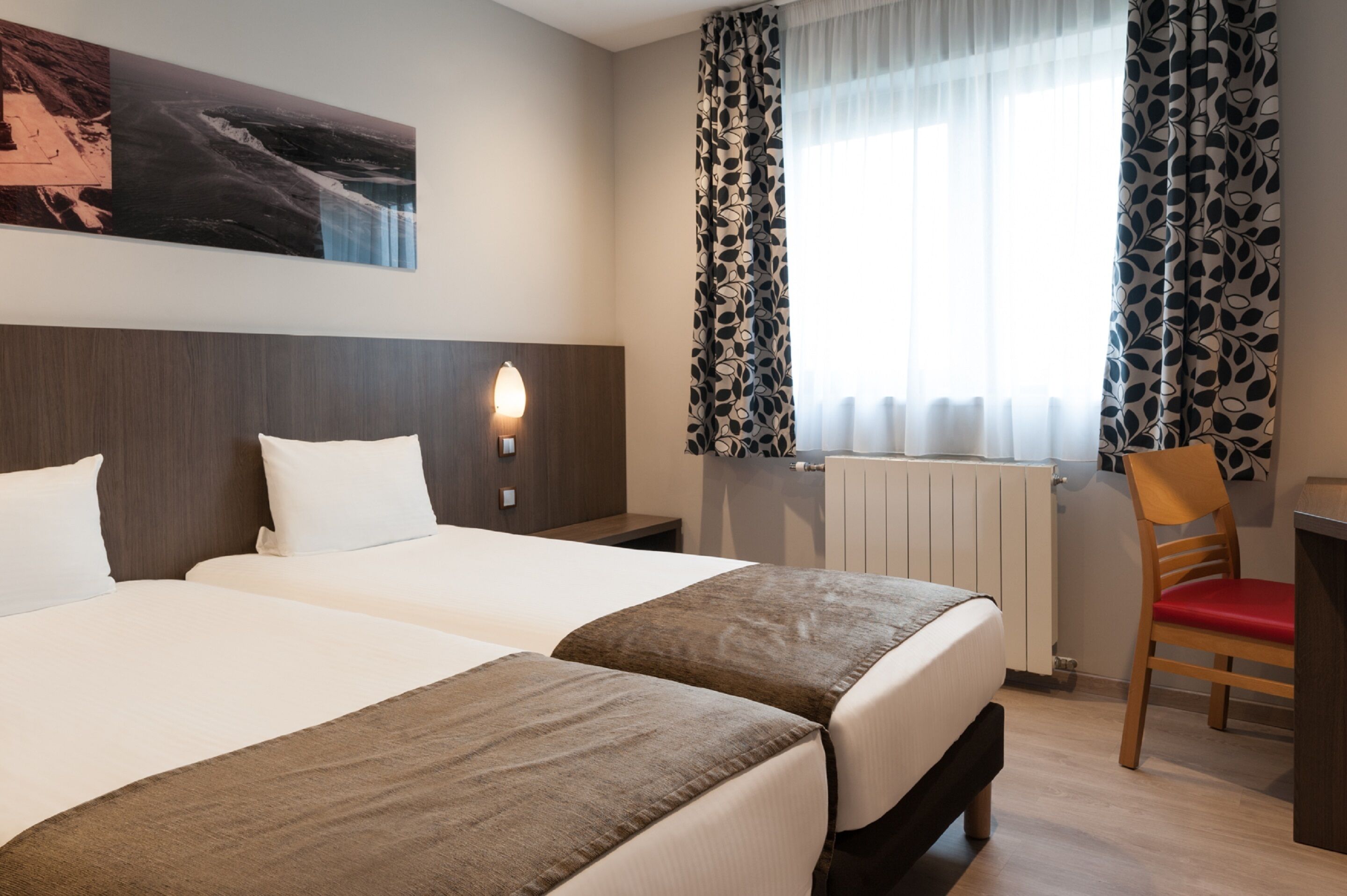 Hotelkamer met twee bedden, een houten hoofdbord, een nachtkastje met een lamp, een raam met gordijnen met patroon, en een stoel.