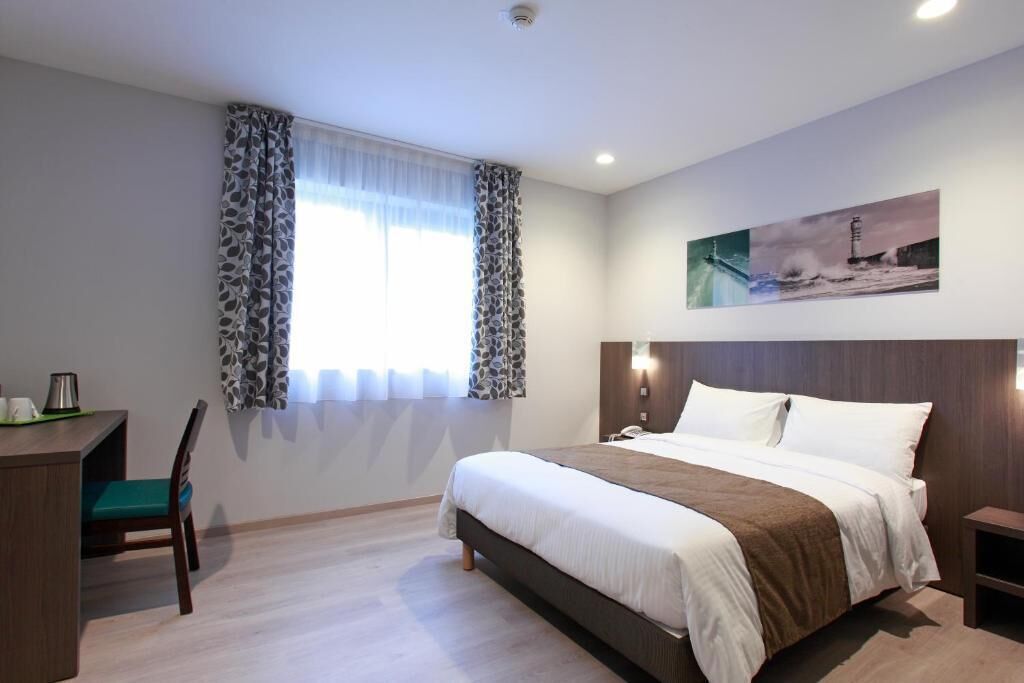 A hotel room with a bed, a desk, a chair, a window with curtains, a painting on the wall, and a nightstand.
