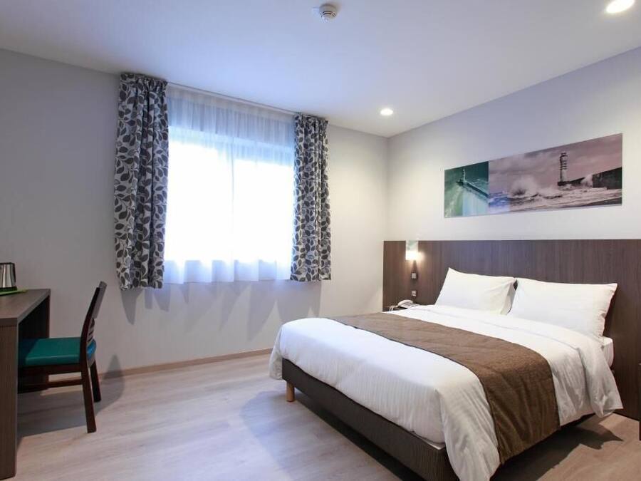 Une chambre d’hôtel avec un lit, un bureau, une chaise, une fenêtre avec des rideaux, un tableau au mur et une table de chevet.