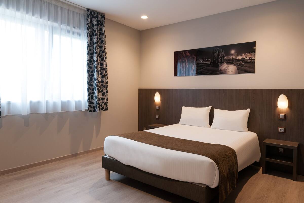 Une chambre d’hôtel avec un grand lit, deux tables de chevet, une petite table d’appoint, une œuvre d’art accrochée au mur et une fenêtre avec des rideaux légers.