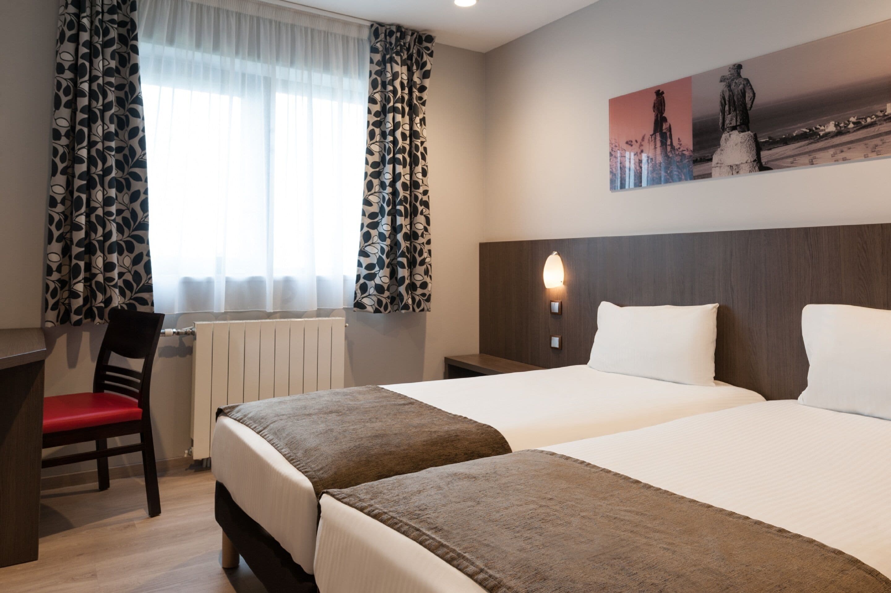A hotel room with two beds, a desk, a chair, and a window with patterned curtains.