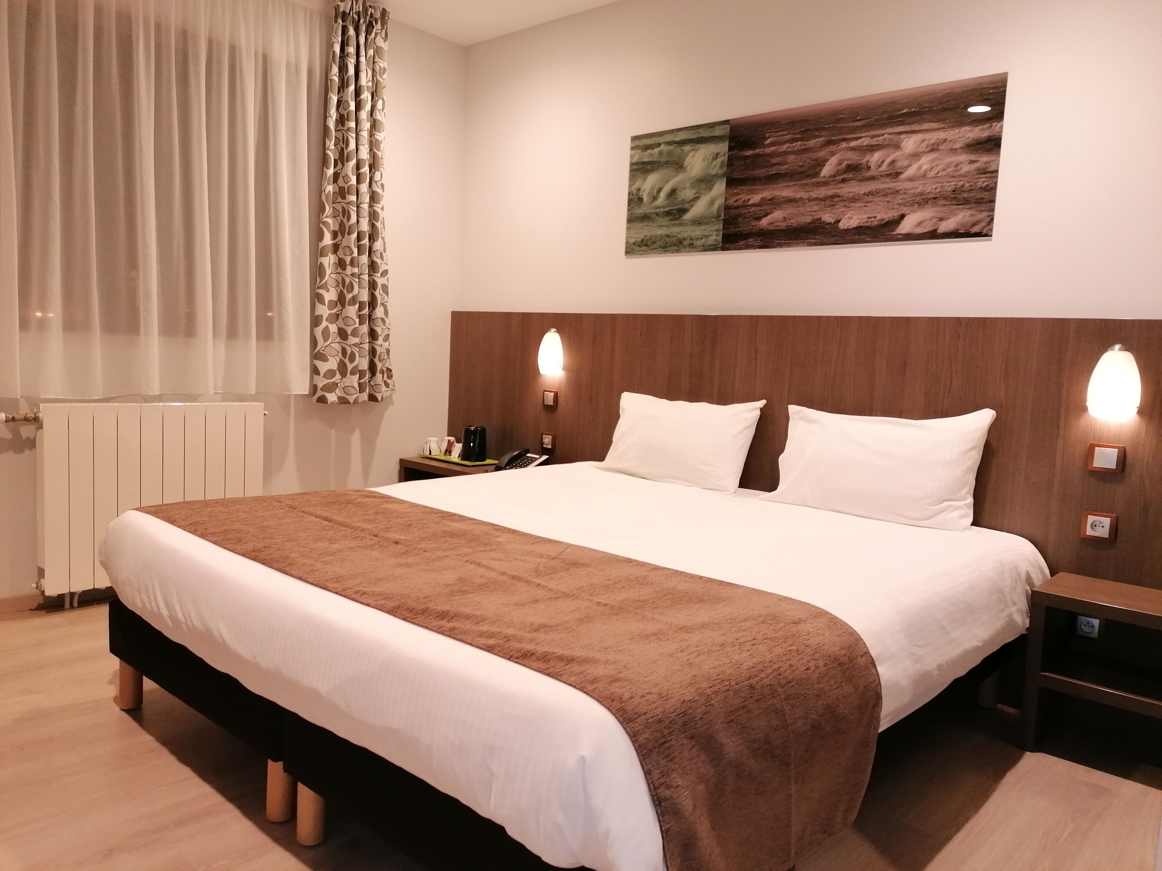 A hotel room with a large bed, a wooden headboard, two bedside lamps, a nightstand, a radiator, and a window with sheer curtains.