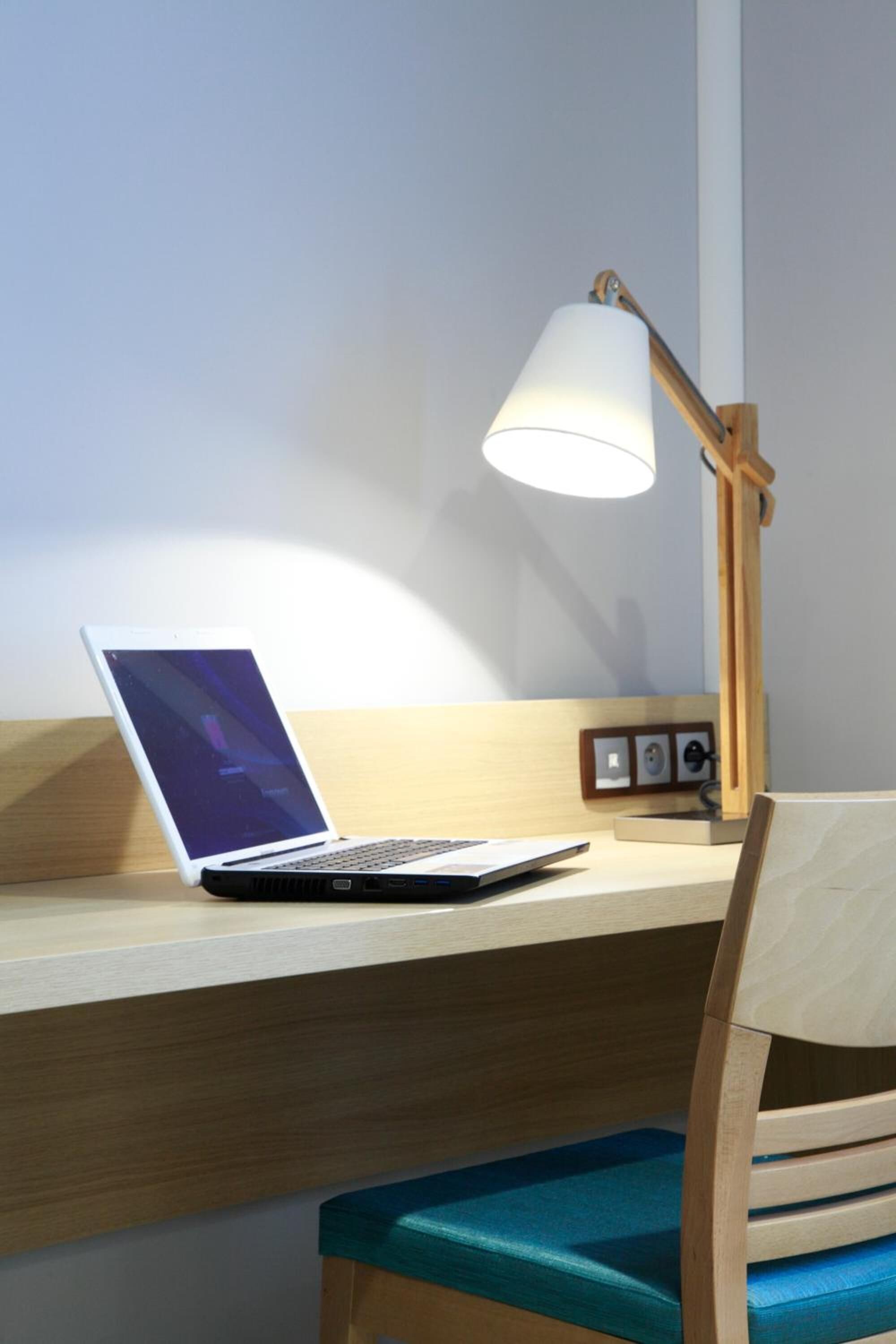 A laptop on a wooden desk with a white lamp and a wooden chair with a blue cushion.