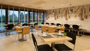 Een moderne eetruimte met meerdere tafels en stoelen, grote ramen met zonwering en een decoratieve wand.