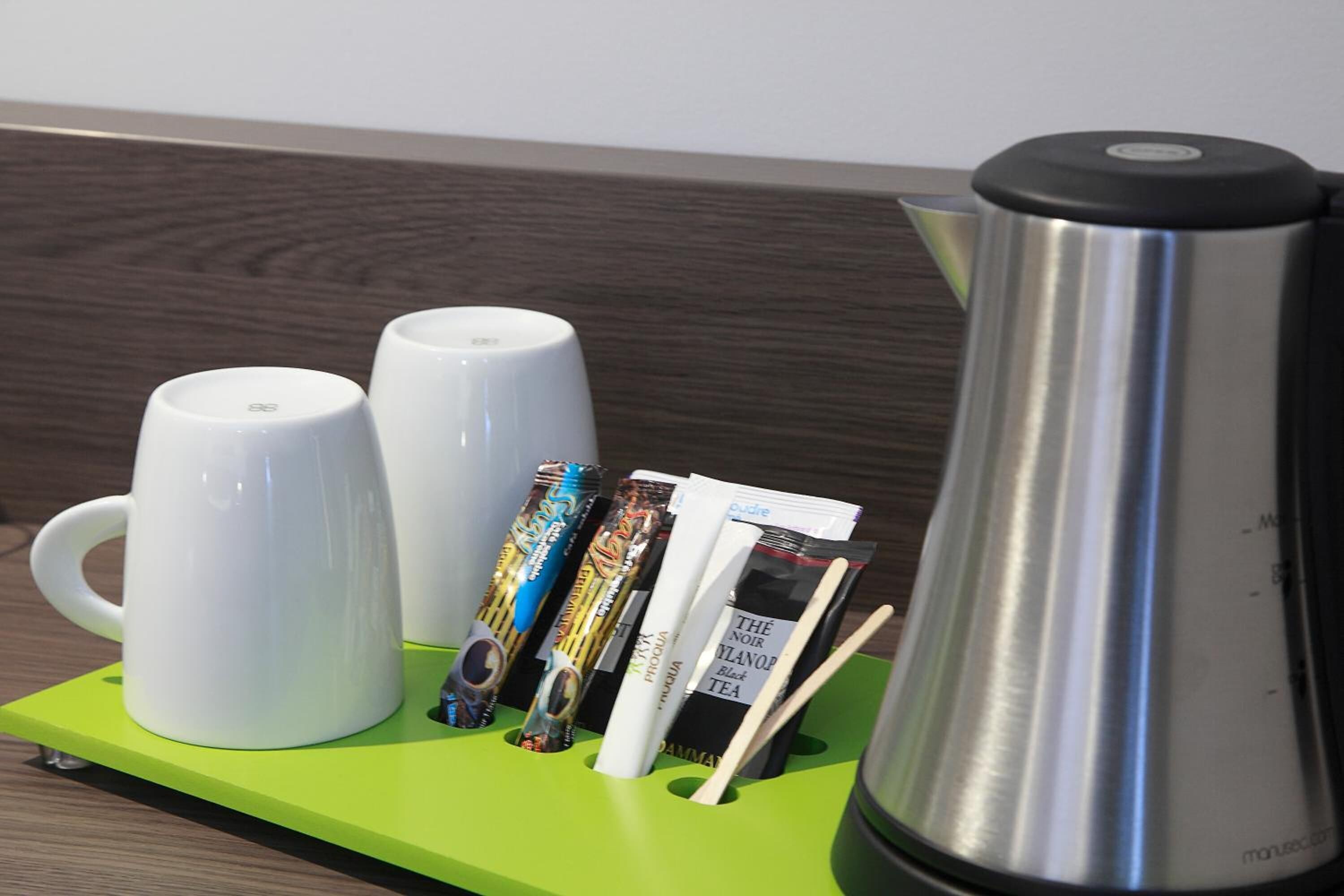 A coffee station with two white mugs, a stainless steel coffee pot, and several coffee packets on a green tray.