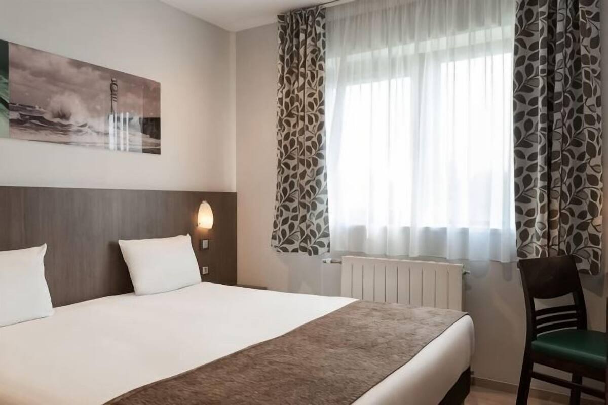 Une chambre d’hôtel avec un lit, une chaise, une lampe fixée au mur, un radiateur et une fenêtre avec des rideaux à motifs.