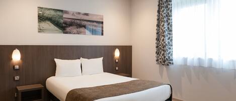 Habitación de hotel moderna con una cama grande, dos mesitas de noche, una lámpara de pared y un ventanal con cortinas translúcidas.