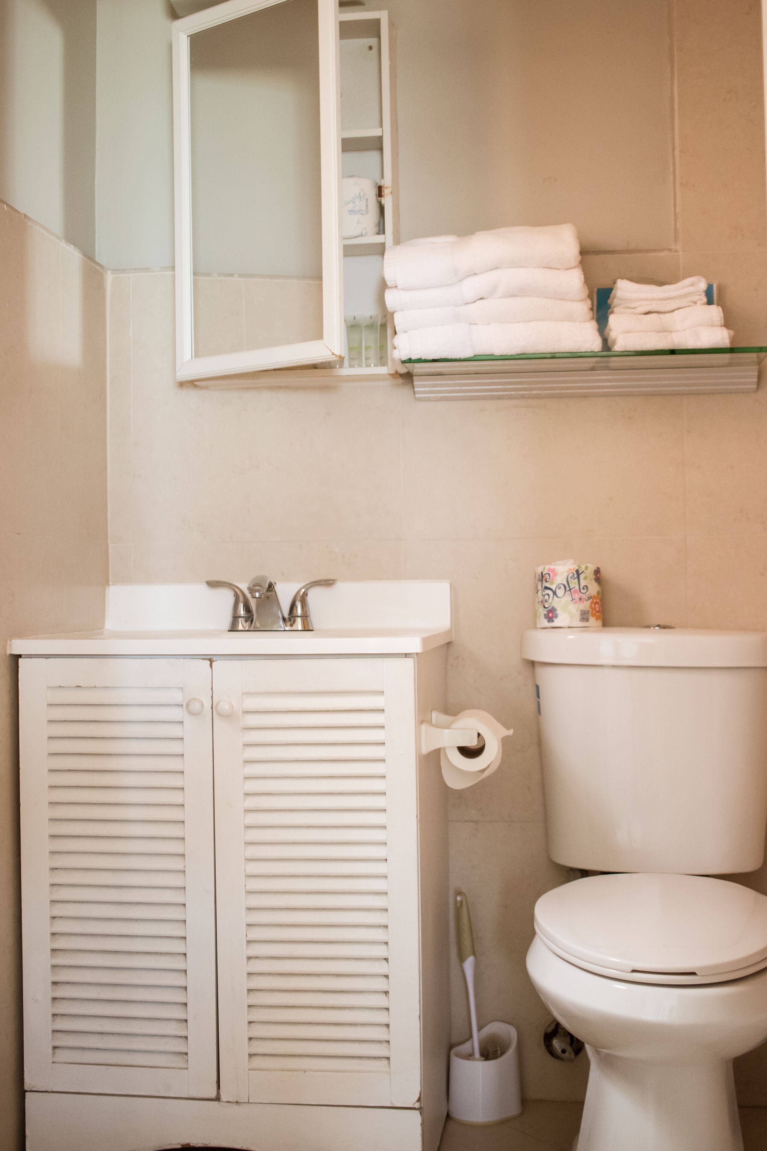 A bathroom with a white sink, toilet, and a mirror above the sink. There are towels stacked on a shelf above the toilet.