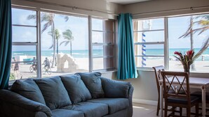 Una habitación con sofá gris, mesa de comedor de madera y sillas, ventanas amplias con cortinas y vista a la playa.