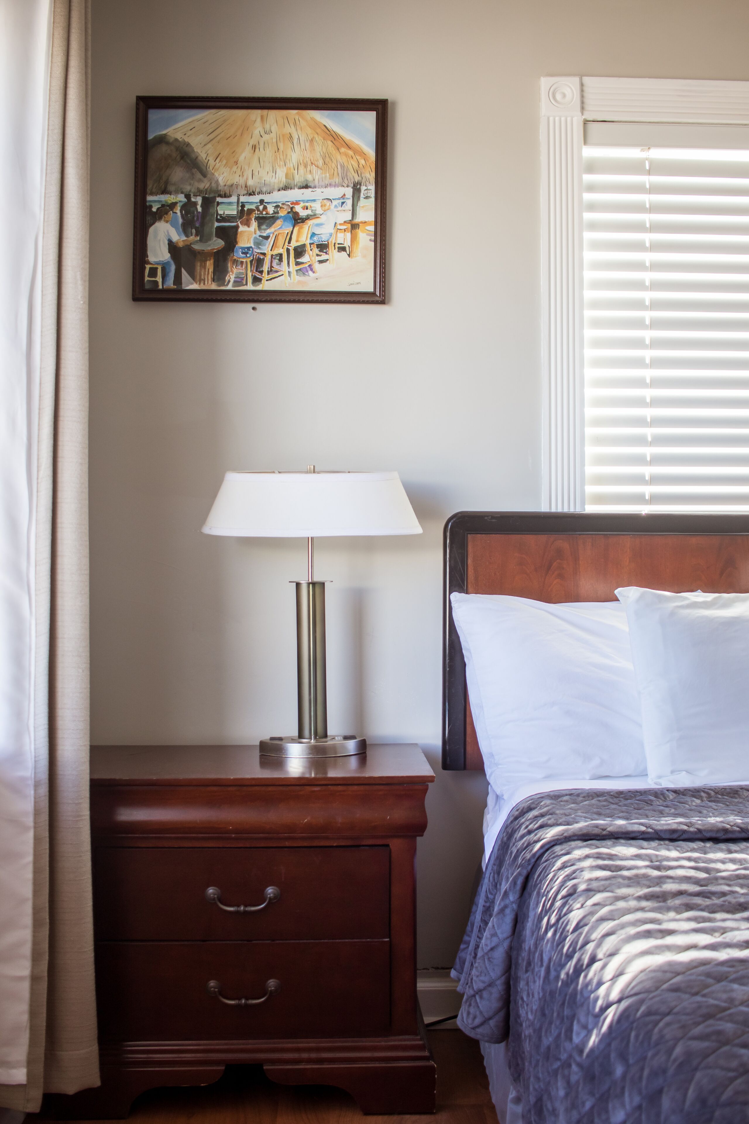A hotel room with a bed, a nightstand with a lamp, a wooden dresser, a painting on the wall, and a window with blinds.