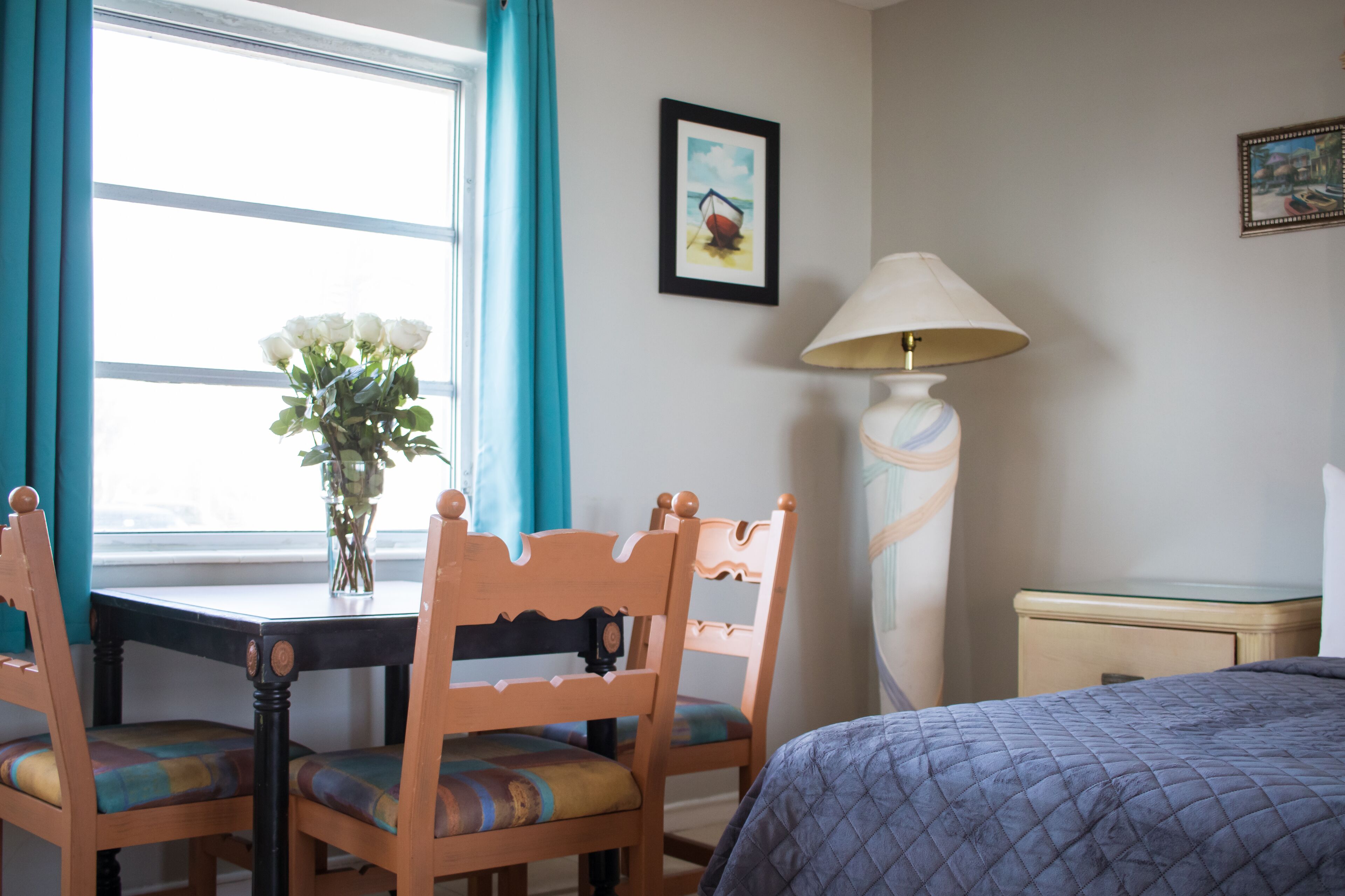 Una habitación de hotel con una cama, una mesa con sillas, un florero con flores, una lámpara y un cuadro en la pared.
