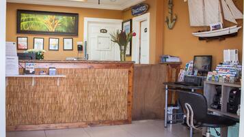 Un área de recepción con un mostrador de madera, una computadora y una silla. Hay un letrero que dice "Beach House" y una fotografía enmarcada de una palmera en la pared.