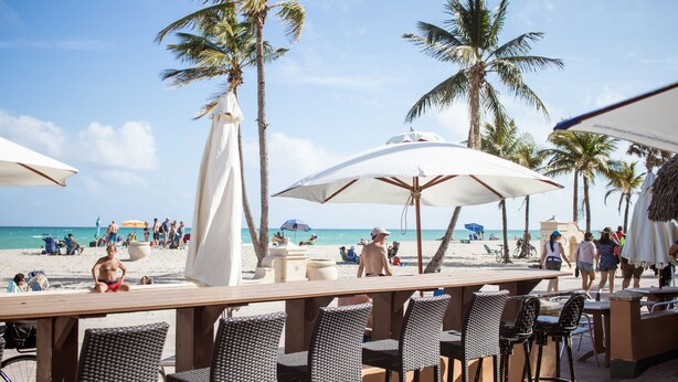 Un bar junto a la playa con mesas al aire libre y sombrillas, con vistas a una playa de arena, palmeras y gente.