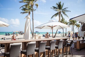 A beachside bar with outdoor seating and umbrellas, overlooking a sandy beach with palm trees and people. - Riptide Oceanfront Hotel (Hollywood)