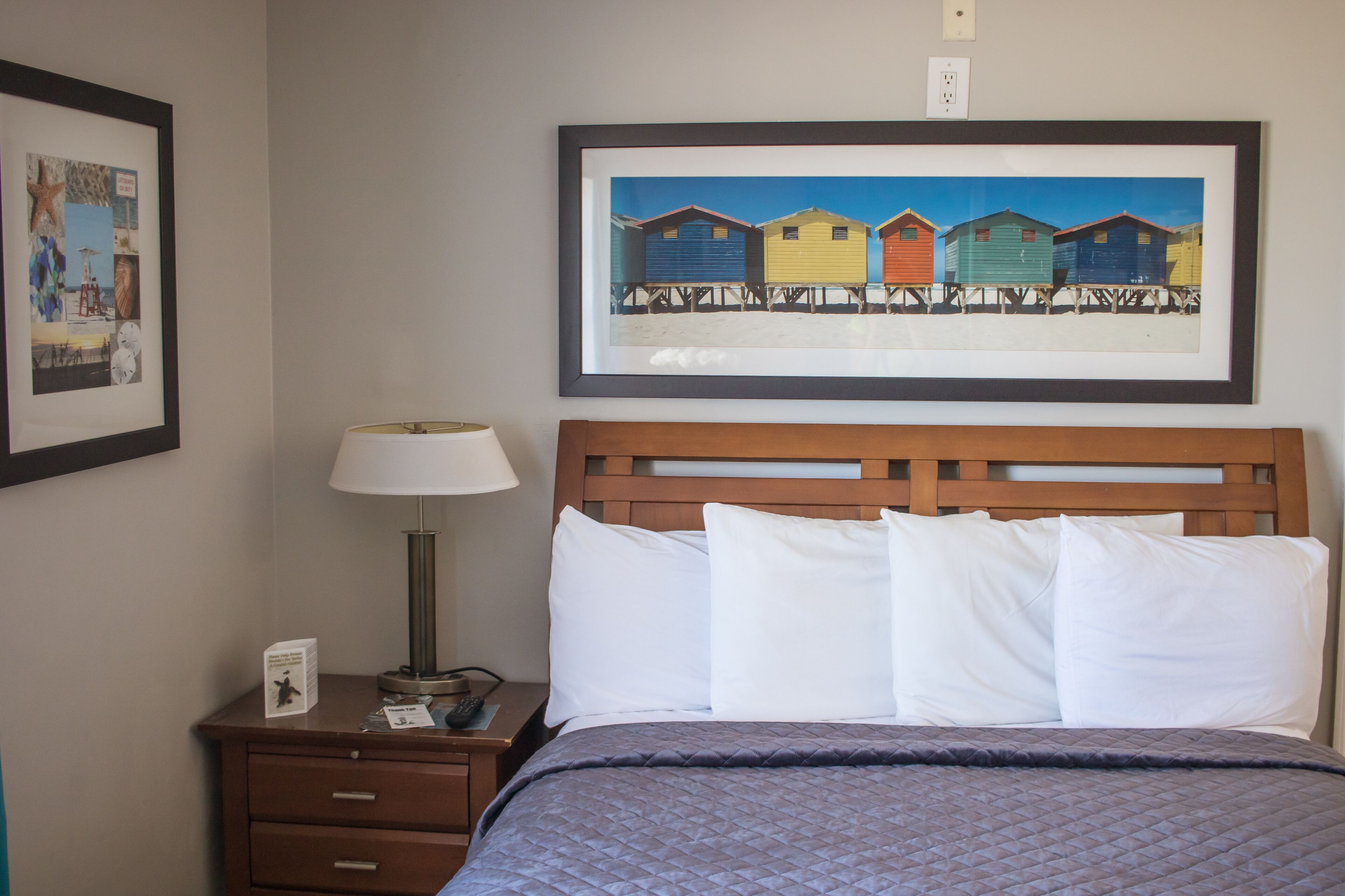 A neatly made bed with white pillows and a quilted blanket, a bedside table with a lamp, and a framed picture of colorful beach houses on the wall.
