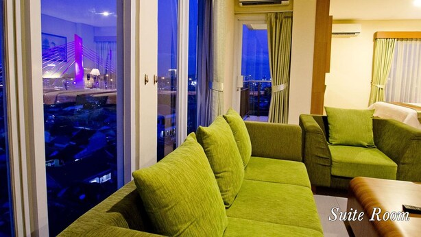 A suite room with a green sofa, a small table, and a view of a cityscape at night. - De Batara Hotel (Bandung)