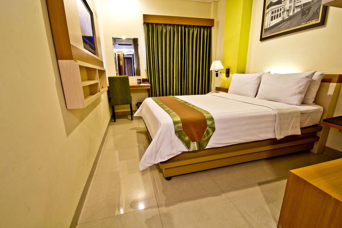 A hotel room with a bed, a desk, a chair, a TV, a mirror, a lamp, and a picture on the wall.