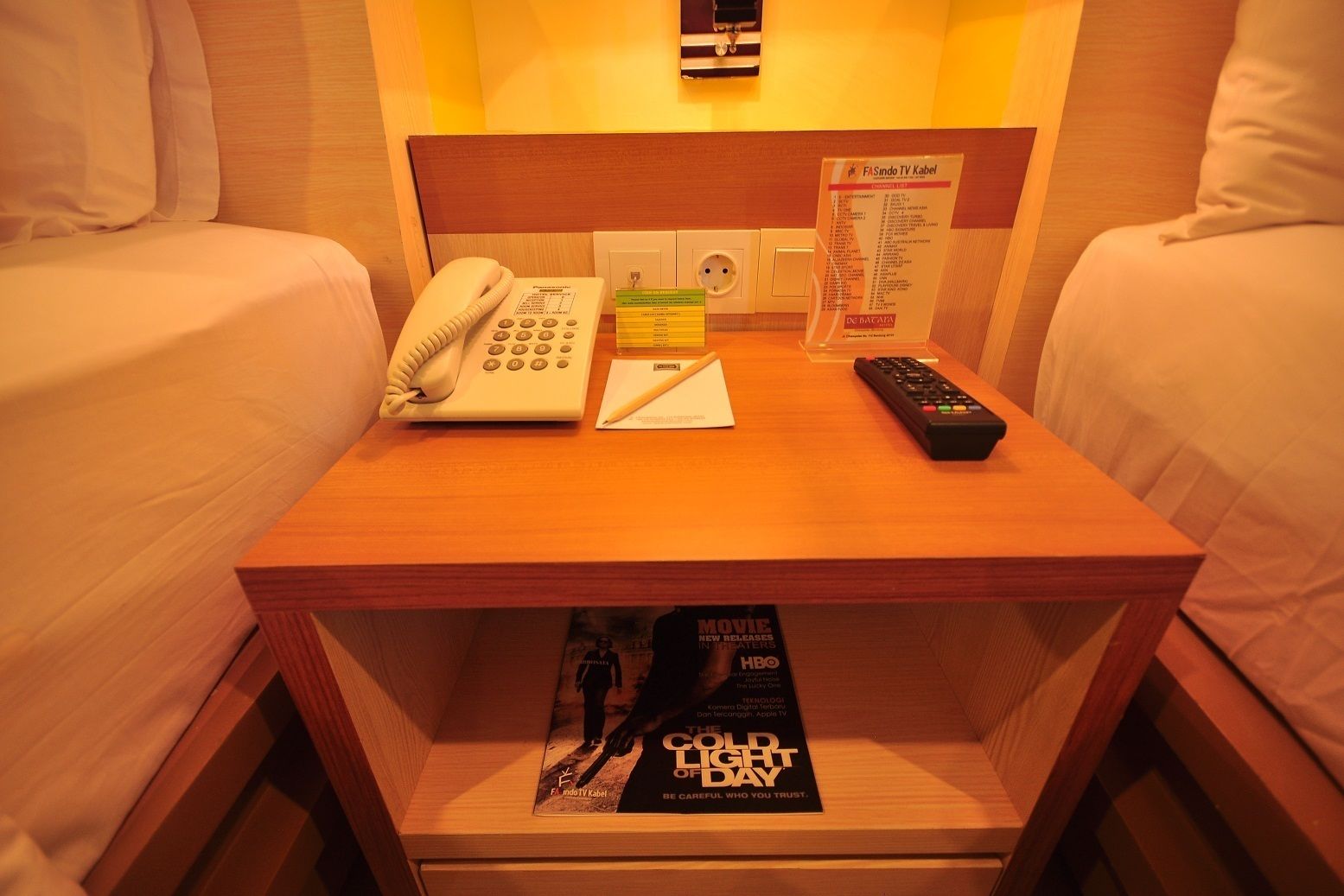 Habitación de hotel con una mesita de noche de madera, un teléfono, un control remoto y una revista sobre la mesa.