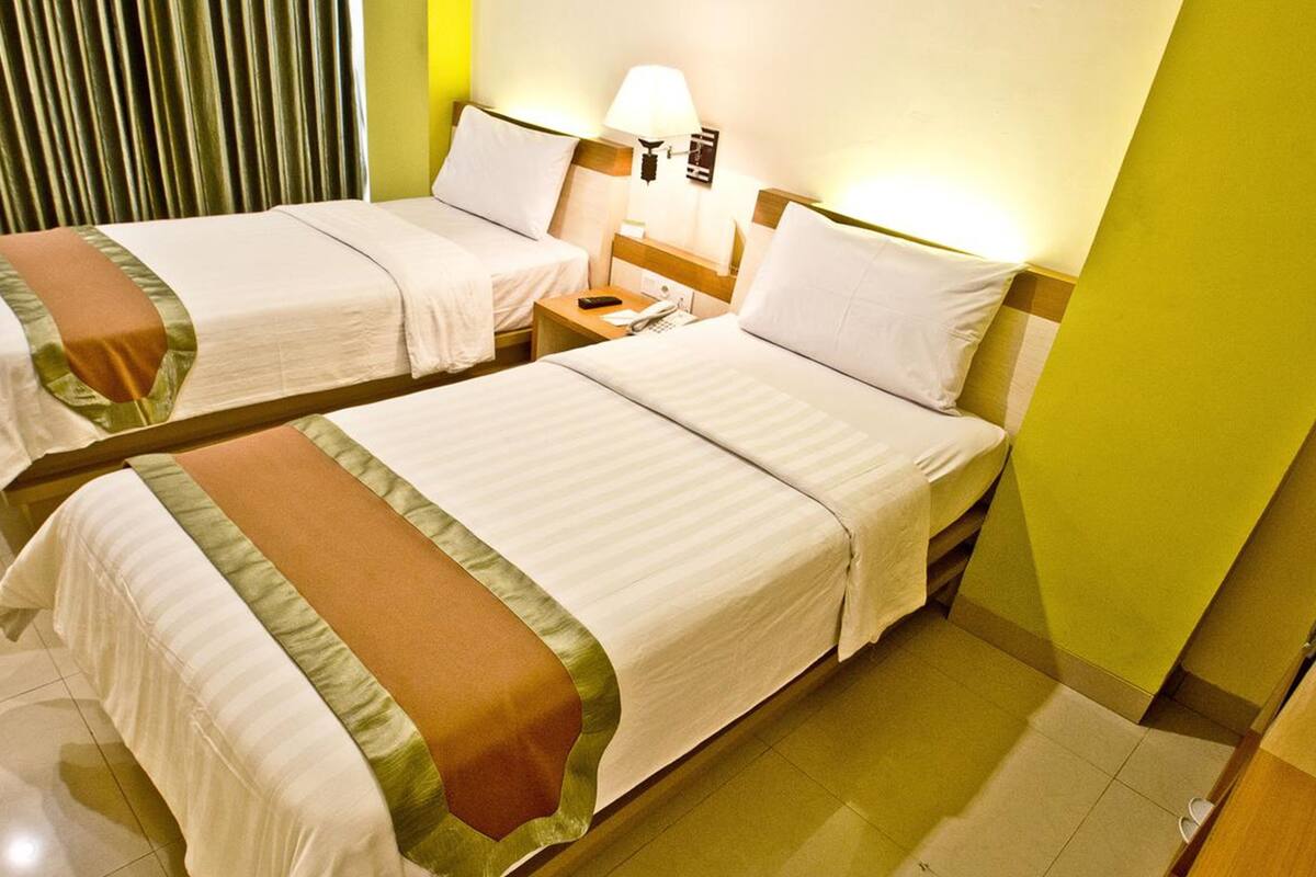Two single beds with white linens and brown decorative borders, a bedside table with a lamp, and a curtain in the background.
