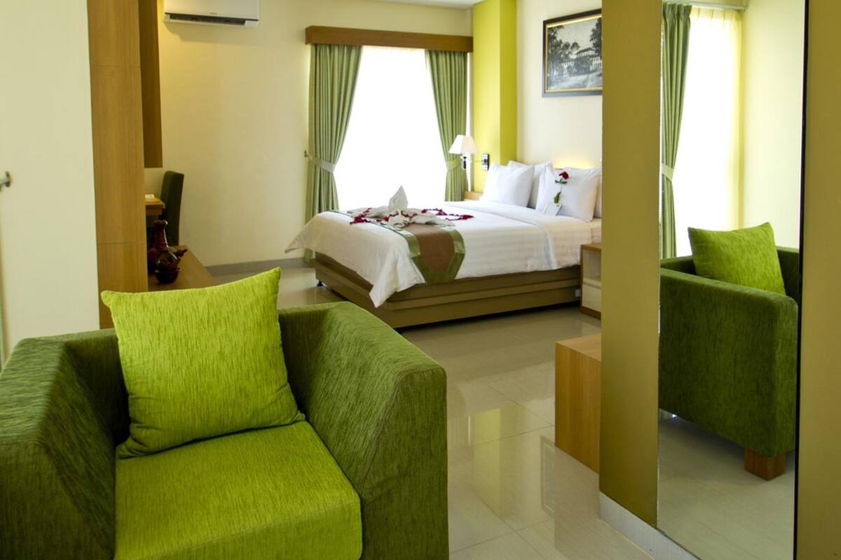 A hotel room with a bed, two green armchairs, a bedside table, and a mirror.