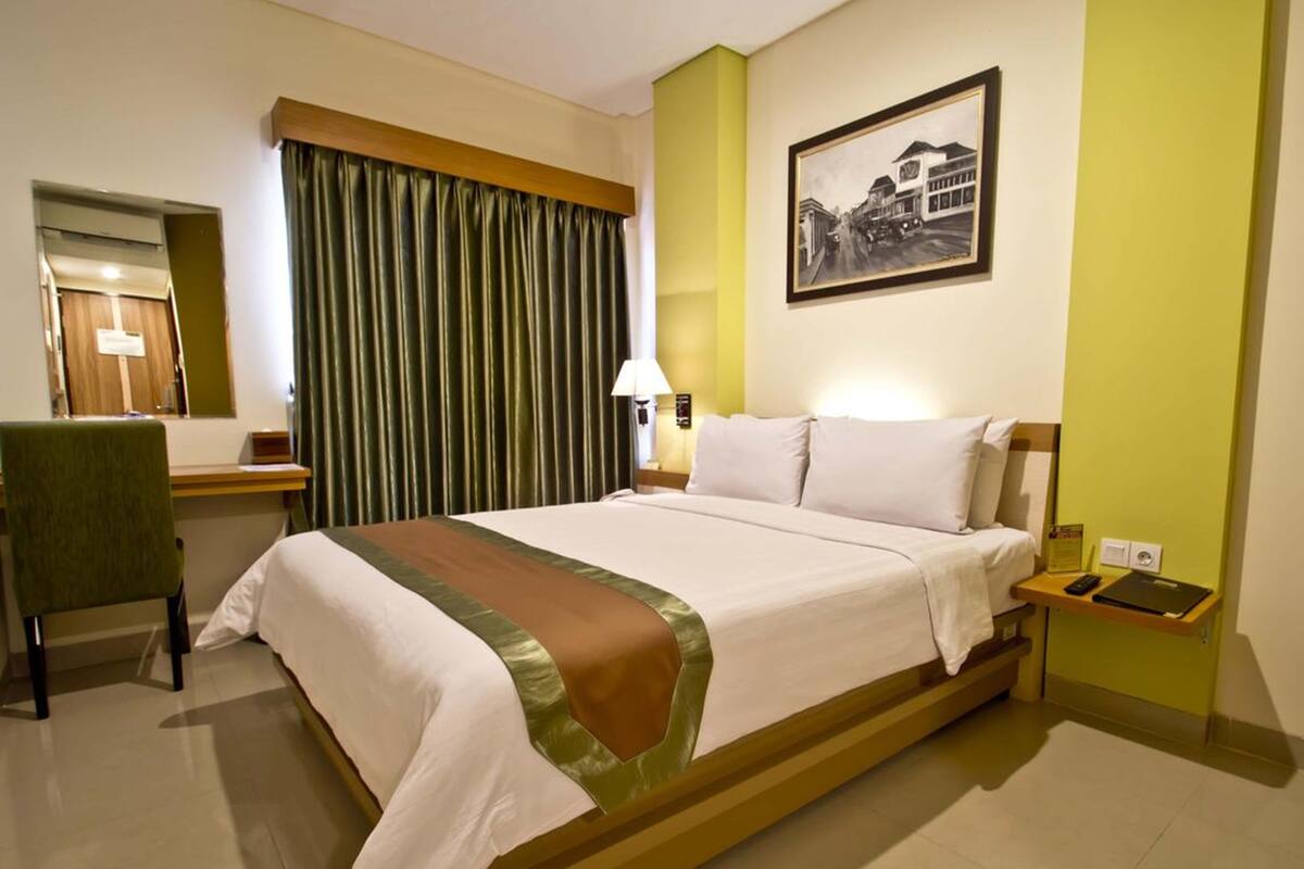 A hotel room with a bed, a desk, a chair, a lamp, and a framed picture on the wall.