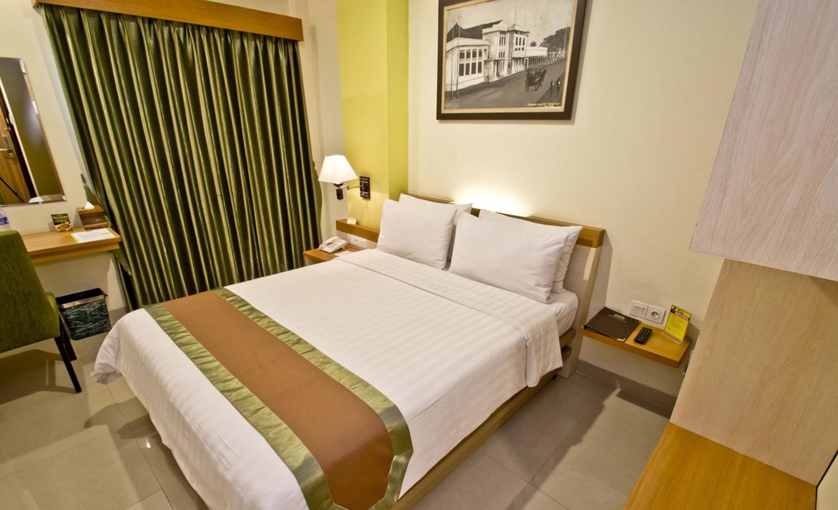 A hotel room with a bed, a desk, a chair, a nightstand, a lamp, a framed picture on the wall, and a trash bin.
