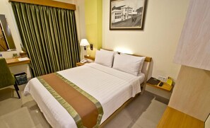Hotellihuone, jossa on sänky, työpöytä, tuoli, yöpöytä, lamppu, seinällä oleva kehystetty kuva ja roskakori.