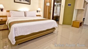 A Deluxe Double Room with a large bed, two bedside tables, a nightstand, a wall-mounted air conditioner, a framed picture, a door to a bathroom, and a small wooden cabinet. - De Batara Hotel (Bandung)