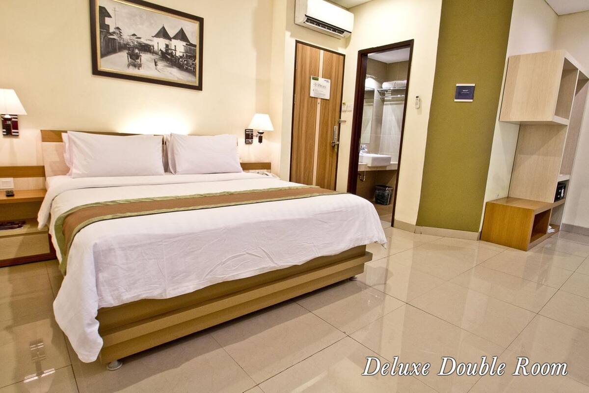 A Deluxe Double Room with a large bed, two bedside tables, a nightstand, a wall-mounted air conditioner, a framed picture, a door to a bathroom, and a small wooden cabinet.