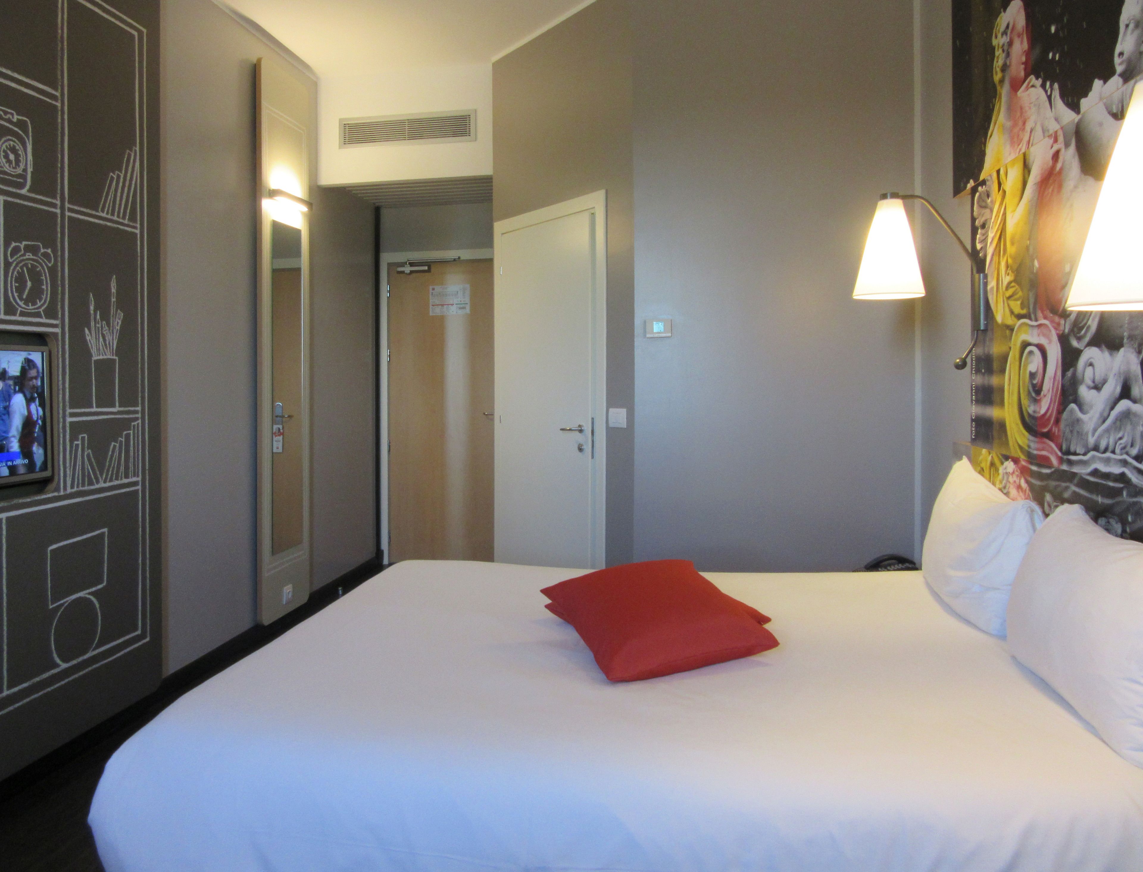 Una camera d'albergo con un letto, un cuscino rosso, una sveglia a parete, una televisione e una porta.