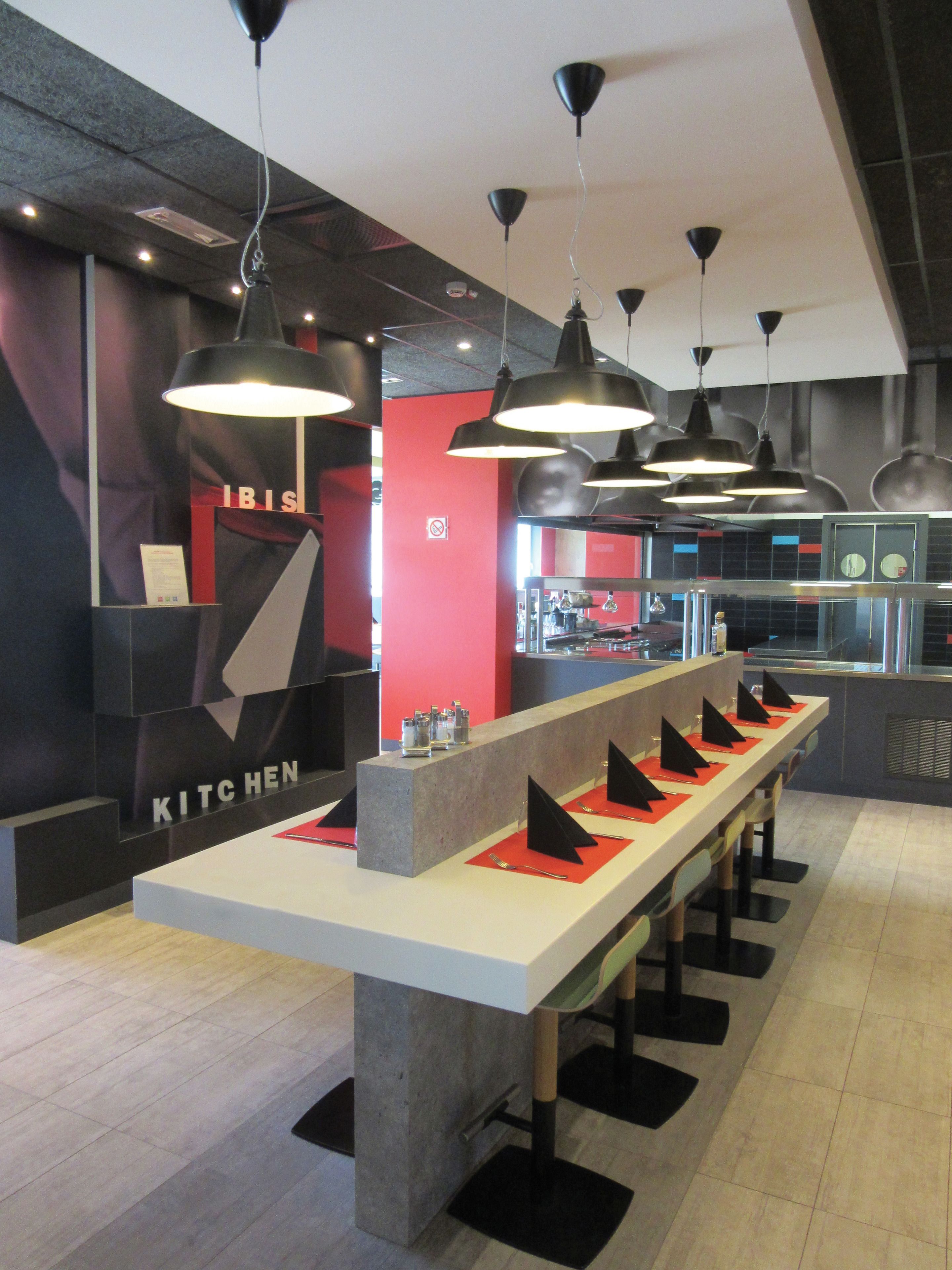 A modern kitchen area with a long counter, multiple black pendant lights, and a red accent wall.