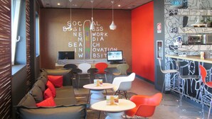 A modern lounge area with a sofa, tables, and chairs, featuring a unique ceiling design with various patterns and lights. - Ibis Milano Fiera (Lainate)