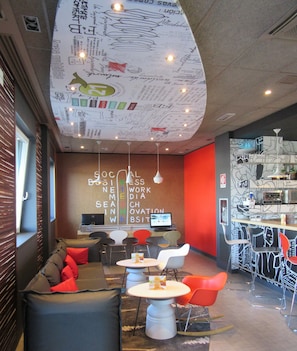 A modern lounge area with a sofa, tables, and chairs, featuring a unique ceiling design with various patterns and lights. - Ibis Milano Fiera (Lainate)