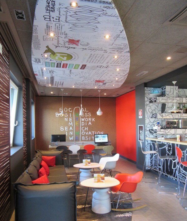 A modern lounge area with a sofa, tables, and chairs, featuring a unique ceiling design with various patterns and lights. - Ibis Milano Fiera (Lainate)