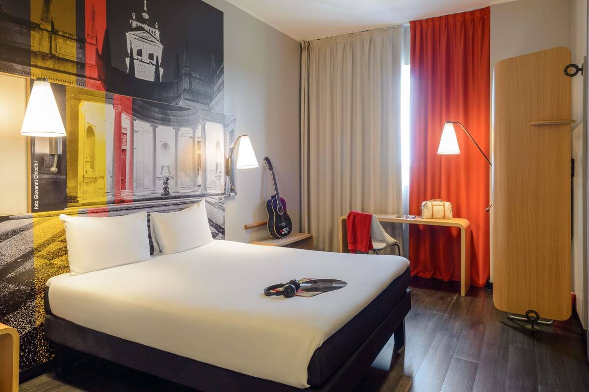 A hotel room with a bed, a desk, a guitar, and a wall with a mural.