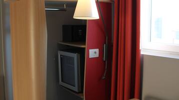 Une pièce de taille modeste, avec une armoire en bois, un petit bureau avec une chaise, une lampe, un four à micro-ondes et une fenêtre ornée de rideaux rouges.