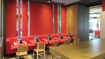 A modern dining area with red seating and wooden tables, featuring a long table in the foreground and a kitchen area in the background.