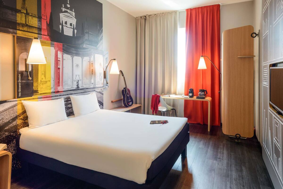 A hotel room with a bed, a desk with a chair, a guitar, a coffee maker, and a TV.
