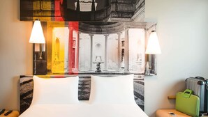 A hotel room with a bed, two bedside lamps, a green suitcase, and a wall-mounted artwork depicting a historical building. - Ibis Milano Fiera (Lainate)