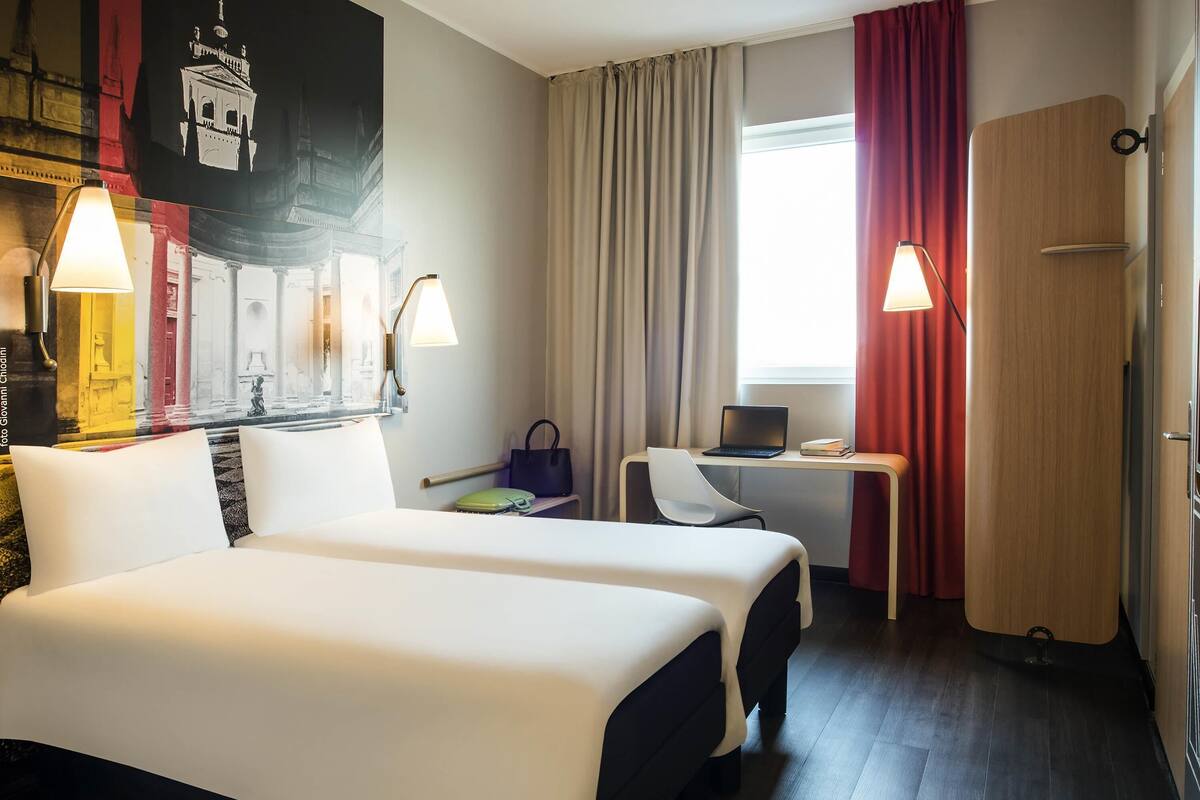A hotel room with two beds, a desk with a laptop, a chair, a lamp, and a window with curtains.