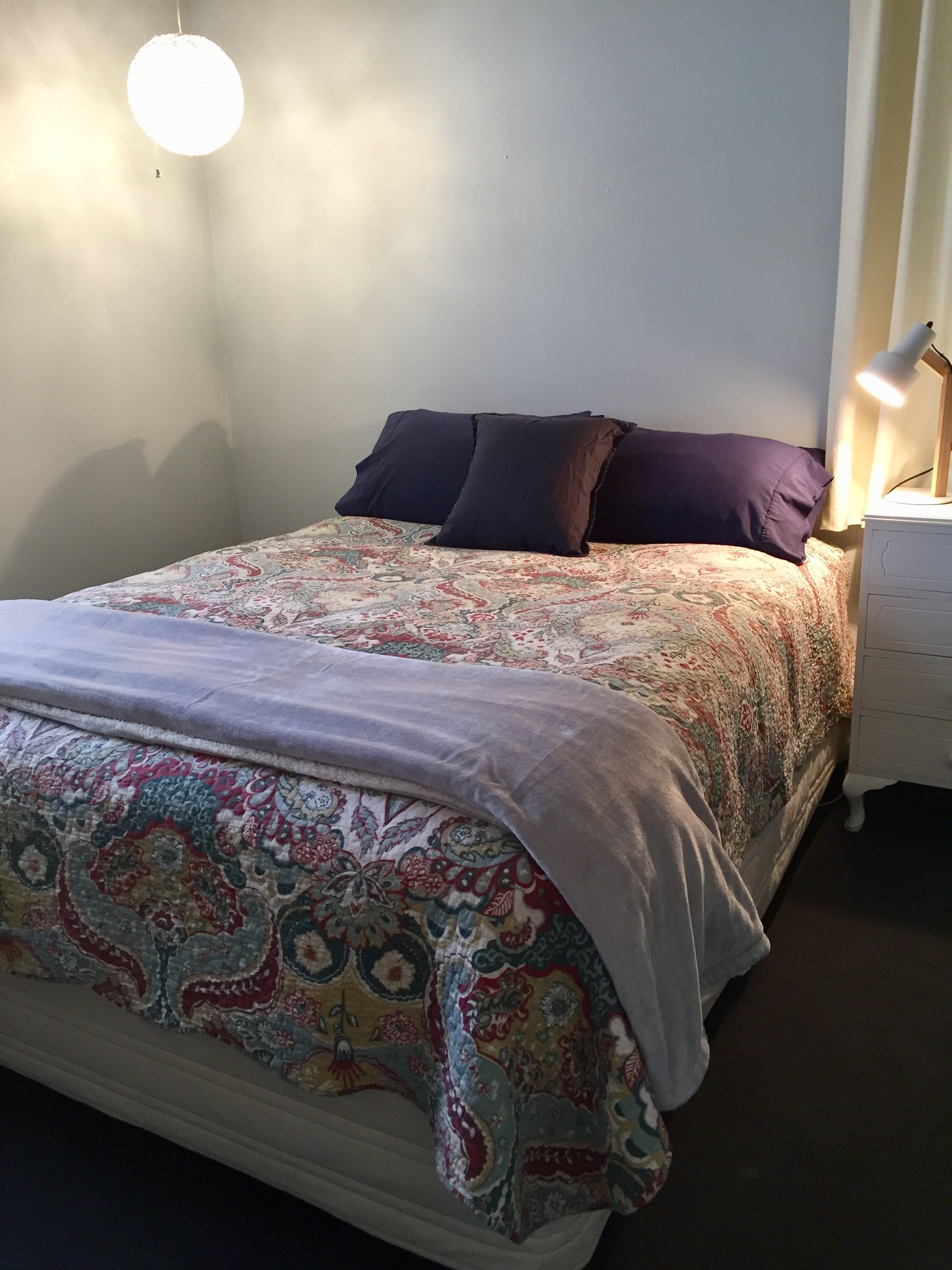 A bed with a patterned bedspread and pillows, a bedside table with a lamp, and a wall-mounted light fixture.