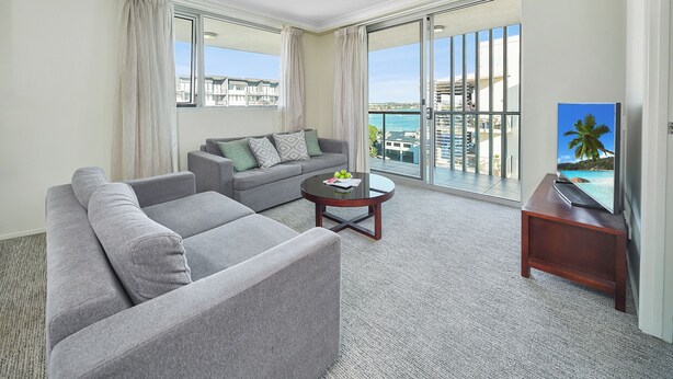 A modern living room with a grey sofa, a smaller grey armchair, and a coffee table. There is a large window with curtains, a flat-screen TV on a wooden stand, and a painting of a palm tree on the wall. - Quest Breakfast Creek (Albion)