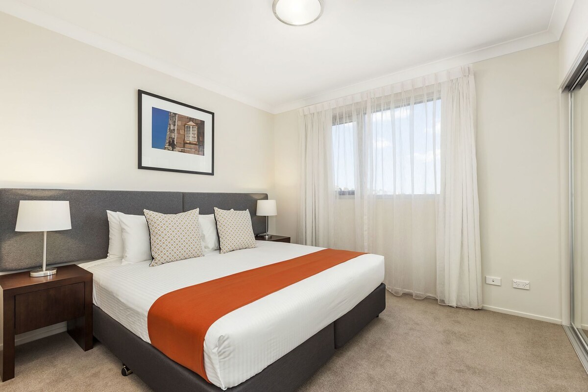 A hotel room with a bed, two bedside tables, a framed picture on the wall, and a window with sheer curtains.