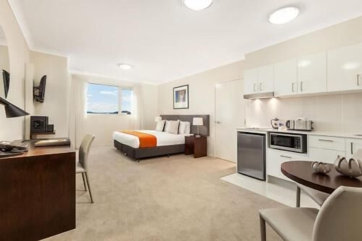 A hotel room with a bed, desk, chair, kitchenette, and a view of the outdoors.