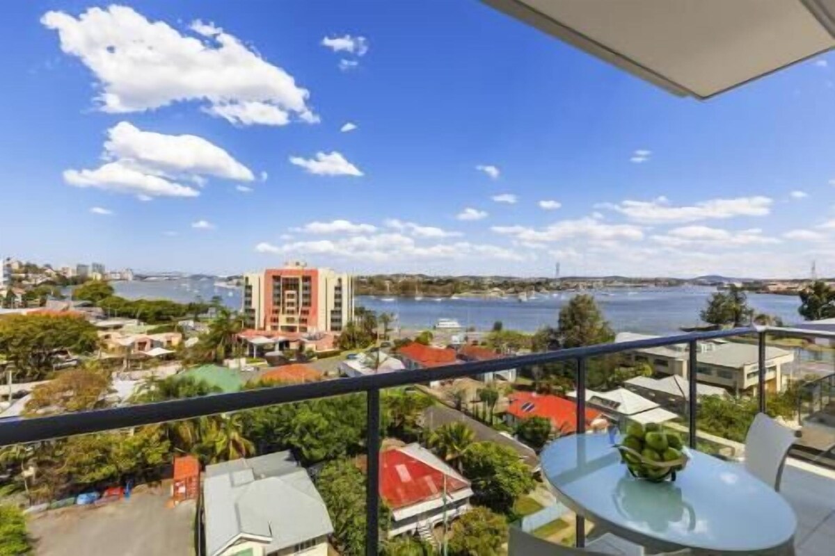 A balcony with a clear view of a body of water, a distant building, and surrounding greenery.